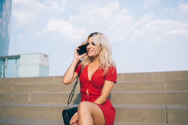 Trendy positive confident lady in red summer dress talking on mobile phone joyfully during rest on city staircase looking away - Fotografie, Obrázek