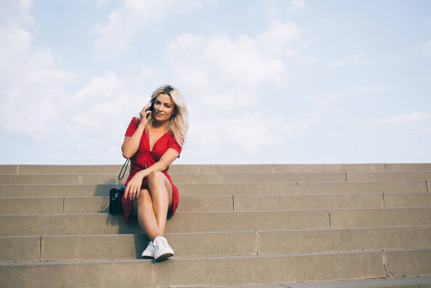 Casual fair hair woman in short red dress sitting on large stone street stairs and chatting on smartphone while looking at camera on background with sky - Fotó, kép
