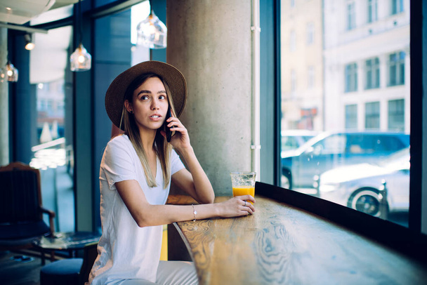 Вид на симпатичную молодую леди со стаканом сока, говорящую по смартфону и смотрящую в сторону, сидя за столом возле окна в ресторане - Фото, изображение