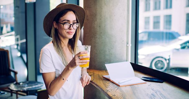 Pensive adult modern female in casual outfit and with glasses wearing hat sitting at high table drinking juice while writing notes against glass window in cafe looking away - Foto, afbeelding