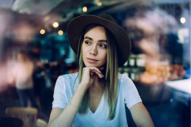 Trough glass view of contemplating young woman in tropical hat and white shirt looking to left side and holding hand near chin while sitting in cafe on blurred background - Valokuva, kuva