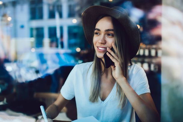 Beautiful and cheerful lady in hat sitting in front of window in cafe and speaking on smartphone while looking away  - Foto, imagen