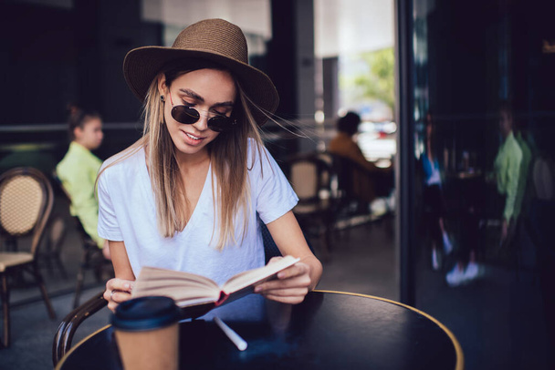 Pleased trendy long haired woman in hat and sunglasses enthusiastically reading book and drinking coffee enjoying rest and waiting order in cafe - Фото, изображение