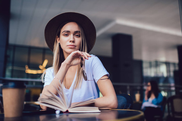 Attractive carefree young woman in stylish hat holding sunglasses and looking at camera while sitting by table with opened book in cafe - Foto, Imagem