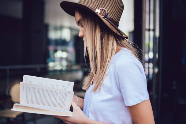 Side view of focused female in hat wearing casual clothes reading book with attention while standing near glass wall on street - Fotografie, Obrázek