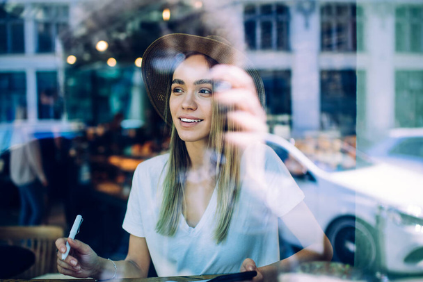 Karkea lasinäkymä hymyilevä nuori nainen trooppisessa hatussa ja valkoisessa paidassa, joka katsoo vasemmalle puolelle istuessaan kahvilassa hämärtyneellä taustalla - Valokuva, kuva