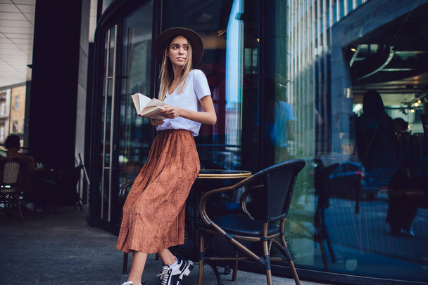 Thoughtful young beautiful woman in hat wearing casual clothes reading book while standing near glass wall and looking away pensively - Foto, Bild