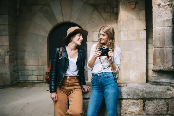 Joyful young attractive woman with blond hair holding camera and looking at cheerful female friend in hat while standing together in street - Фото, зображення