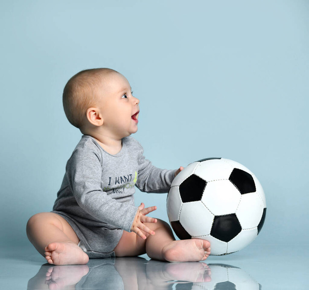 Ginger child in gray bodysuit, barefoot. He holding soccer ball, sitting on floor against blue background. Close up, copy space - Fotó, kép