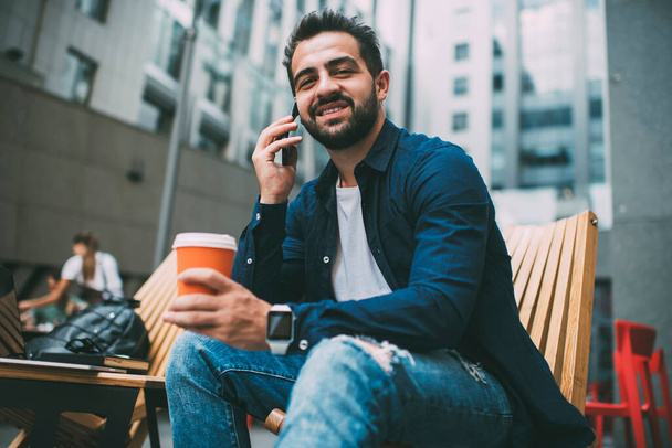 Handsome caucasian male student recreating during coffee break talking on mobile phone in roaming, portrait of young prosperous hipster guy sitting outdoors enjoying smartphone conversation  - Zdjęcie, obraz