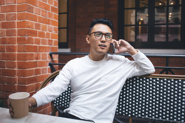Focused young ethnic man in glasses looking away and speaking on phone while holding takeaway coffee and relaxing at table outdoors - Фото, изображение