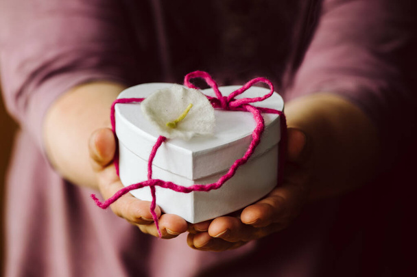 Mani che tengono una piccola scatola regalo bianca a forma di cuore con un fiocco rosa autofatto e un fiore. profondità di campo poco profonda
 - Foto, immagini