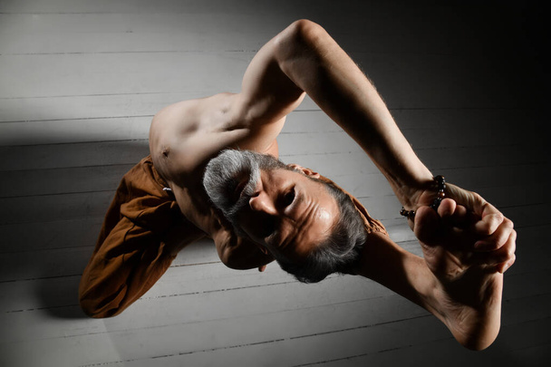 Aged yogi man with naked torso, in brown pants and bracelets is performing his flexibility. Lying on a white wooden floor - Valokuva, kuva