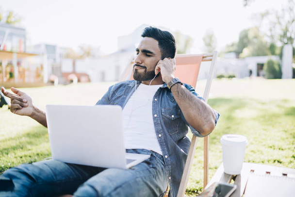 Smart relaxed smiling young ethnic male in casual denim shirt listening to music with laptop and earphones spending time in sunny park - Foto, Imagen