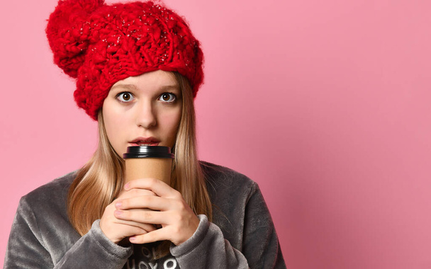Teenage girl in gray pajamas, red hat. She is holding a paper cup and going to drink, posing on pink background. Close up - Foto, Imagen