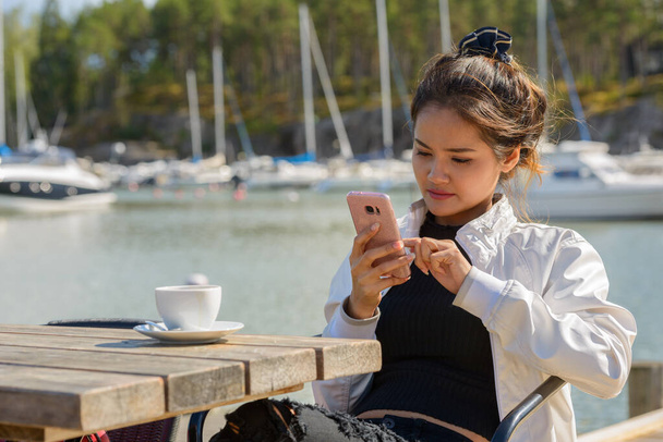 Young beautiful Asian tourist woman using phone at restaurant by the pier - Photo, Image