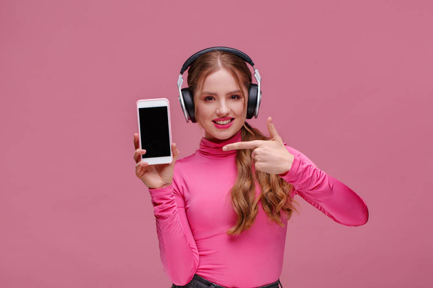 Funny redhead girl showing blank screen mobile phone on camera. Young smiling woman wearing earphones demonstrating smartphone display isolated over pink background. Copy space. App for your business - Photo, image