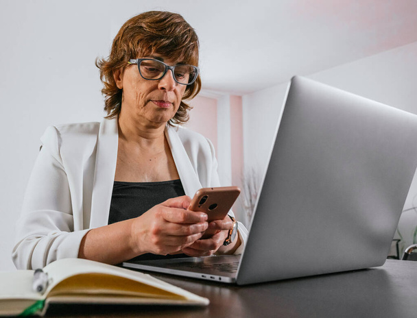 Old lady using her smartphone while working on her laptop. Business woman working from home. Senior woman working from home. Female entrepreneur works from home. Senior woman entrepreneur. - Photo, Image