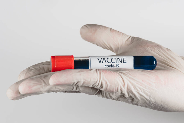 Tubo de vacío con una muestra de vacuna Covid-19 de color azul en la mano con un guante médico sobre fondo blanco
 - Foto, Imagen