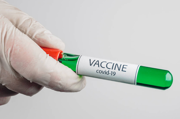 Tubo de vacío con una muestra de vacuna Covid-19 de color verde en la mano con un guante médico sobre fondo blanco
 - Foto, Imagen