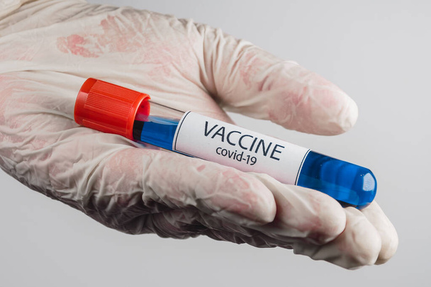 Elinde mavi renkli Covid-19 aşısı olan bir vakum tüpü ve beyaz arka planda tıbbi bir eldiven. - Fotoğraf, Görsel