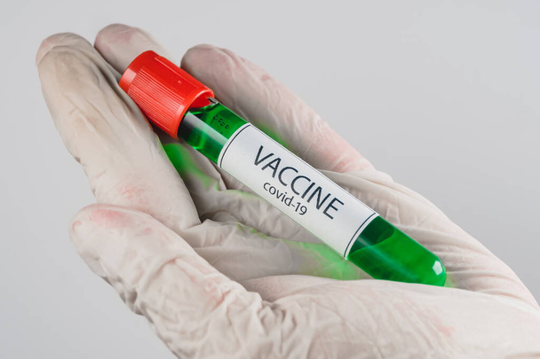 Tubo de vacío con una muestra de vacuna Covid-19 de color verde en la mano con un guante médico sobre fondo blanco
 - Foto, Imagen