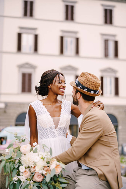 Multiracial wedding couple. Wedding in Florence, Italy. African-American bride in a white dress with a long veil and a bouquet, and Caucasian groom in a sandy jacket and straw hat. - Photo, Image