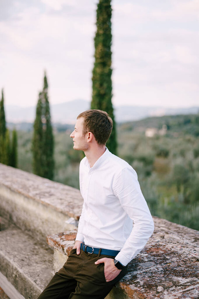 Un grand portrait masculin sur le toit d'une ancienne villa viticole en Toscane, Italie. Chemise blanche à manches longues, montres intelligentes, ceinture bleue, mains dans les poches de pantalon
. - Photo, image
