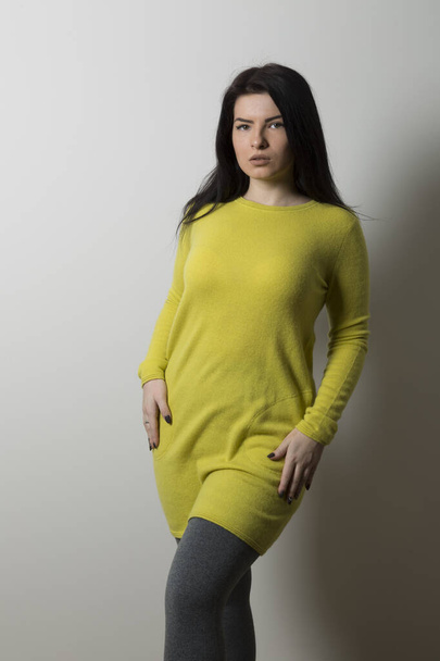 A girl with a yellow long sweater and gray pockets in gray leggings stands on a gray wall background - Фото, изображение