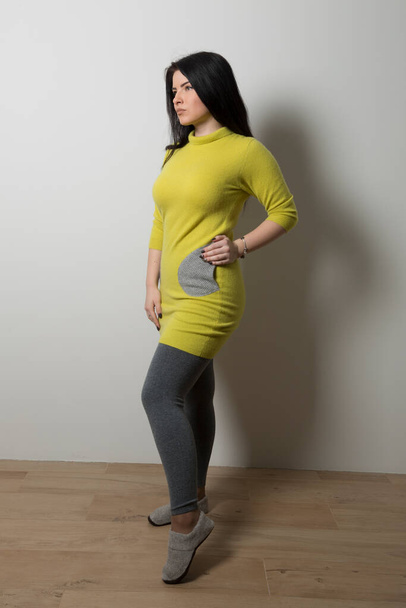 A girl with a yellow long sweater and gray pockets in gray leggings stands on a gray wall background - Foto, afbeelding