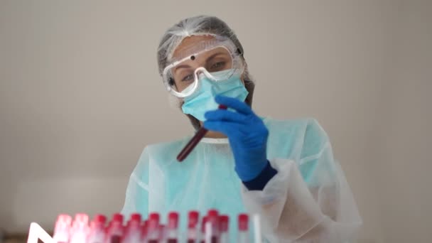Girl laboratory assistant in a protective suit, mask and gloves holds a test tube with a blood sample infected with coronovirus Covid-19. Diagnostics of sars-cov-2 - Кадри, відео