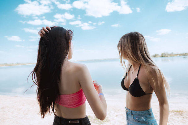 Δύο νεαρά κορίτσια στέκονται στην παραλία και βλέπουν προς τη θάλασσα μια ζεστή ηλιόλουστη μέρα. - Φωτογραφία, εικόνα