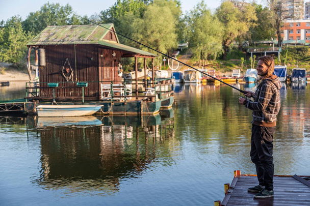 長い髪の男は筏から川の角で釣りをしている。ボートや浮動小数点の家を背景に木で彼の周りにあります。回転リール、緑のラインとルアーと釣竿を使用しています。. - 写真・画像