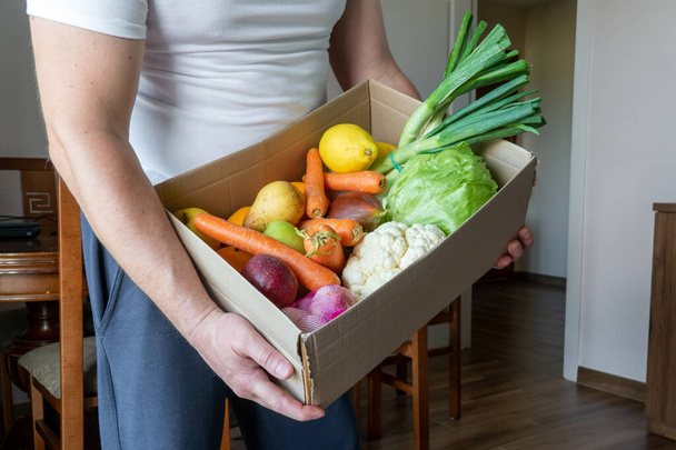 Egy önkéntes, egy ételszállító egy doboz zöldséget tart a kezében. Zöldségek egy kartondobozban, közelkép. Friss zöldségek kartondobozban barna fa háttérrel. Vegetáriánus doboz. - Fotó, kép
