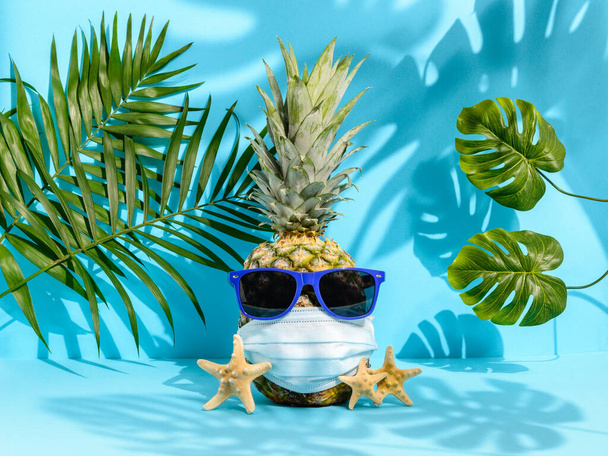 Piña en gafas de sol y máscara médica entre hojas de palma y monstera sobre fondo azul. Fuertes sombras tropicales. Vacaciones hawaianas en el paraíso durante la pandemia de Coronavirus. Concepto verano 2020
 - Foto, imagen