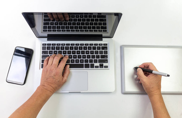 Воздушный снимок рук, работающих с ноутбуком и графическим планшетом с мобильным телефоном в стороне на белом столе
 - Фото, изображение