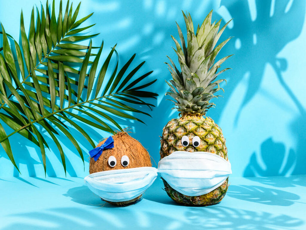 Roztomilý ovocný pár kokos a ananas s očima v lékařských maskách mezi palmovými listy na modrém pozadí. Silné tropické stíny. Havajská dovolená během pandemie koronaviru. Koncept léta 2020 - Fotografie, Obrázek