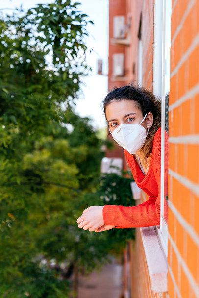 Teenager v masce, dívající se z okna. Společenská vzdálenost. Koronavirus. Dospívající dívka zamčená ve svém domě kvůli pandemii. Smutný teenager. Mladá dívka s ochrannou maskou - Fotografie, Obrázek