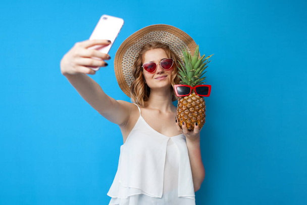 young girl in a sun hat and glasses takes a selfie with pineapple on a blue isolated background, woman tourist taking pictures on vacation - Foto, Imagen