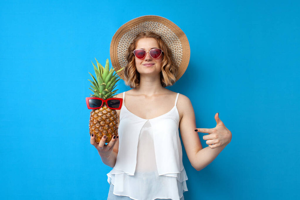 niña en un sombrero de sol y gafas muestra un dedo en una piña sobre un fondo azul aislado, turista mujer en un complejo, concepto de vacaciones de verano
 - Foto, Imagen
