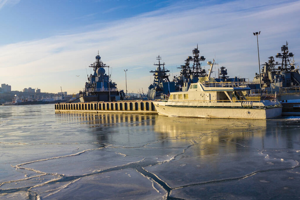 Военные эсминцы, флотилия стоит на дороге в морском порту. Холодное время года вода покрыта тонким слоем льда. Вдалеке очертания города. Доброе утро, солнечный свет
. - Фото, изображение