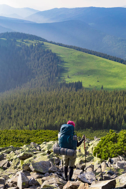 Hikers girl is walking in mountains. Young woman with a tourist backpack and trekking poles walks down a mountain slope dotted with large stones against the backdrop of mountain slopes and a forest. Hiking in the mountains. - Φωτογραφία, εικόνα