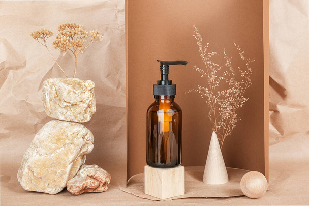 Brown glass bottle with pump of cosmetic products on wooden geometric shapes, stones and dried flowers on beige paper background. Natural Organic Spa Cosmetic Beauty concept Front view Mockup. - Фото, зображення