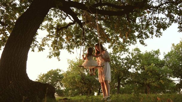 Mom shakes a healthy little daughter on swing under tree in sun. mother plays with child they are swinging on a rope on an oak branch in forest. girl laughs, rejoices. Free family having fun in park - Photo, Image