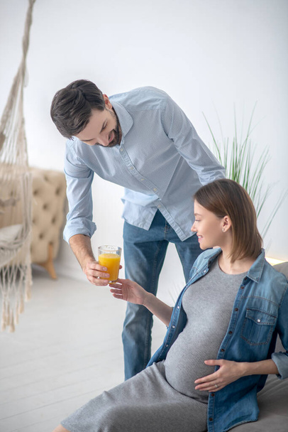 Dark-haired man giving a glass of orange juice to his pregnant wife and smiling - Photo, Image