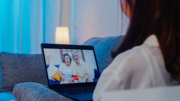 Ázsiai diák csere nő segítségével laptop video hívás beszél a családdal, miközben dolgozik otthonról a nappaliban éjszaka. Önizoláció, társadalmi távolság, karantén a következő normális esetben. - Fotó, kép