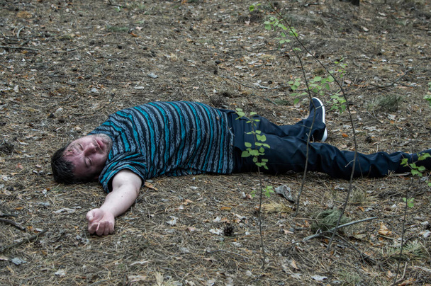 Asesinato en el bosque. El cuerpo de un hombre con una camiseta y pantalones azules yace en el suelo entre los árboles del bosque. Víctima de un ataque. Fotografía horizontal. - Foto, Imagen