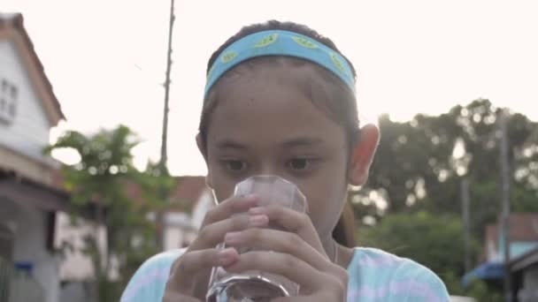 Υγιές ασιατικό χαριτωμένο κορίτσι κρατώντας ένα ποτήρι και πίνουν μεταλλικό νερό μετά την άσκηση. Όμορφη έφηβη με ρούχα γυμναστικής πίνει ένα ποτήρι νερό ενώ στέκεται μπροστά στο σπίτι της.. - Πλάνα, βίντεο
