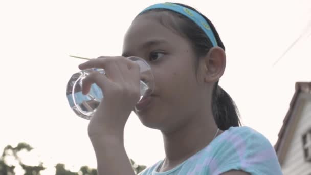 Saudável asiático bonito menina segurando um copo e beber água mineral após o exercício. Adolescente bonita em roupas de exercício está bebendo um copo de água enquanto está em frente a sua casa
. - Filmagem, Vídeo