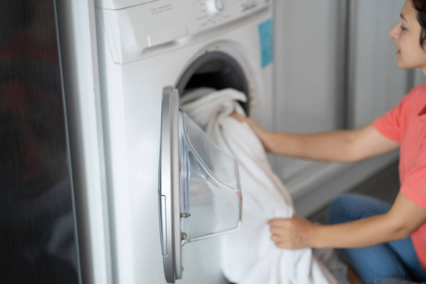 A girl loads dirty laundry into a washing machine while sitting on the floor in an apartment. Laundry day, housework. - Valokuva, kuva